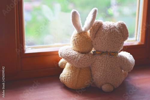 Photographie Best friends teddy bear and bunny toy sitting on brown window sill hugging each other and looking out of window on vintage tone