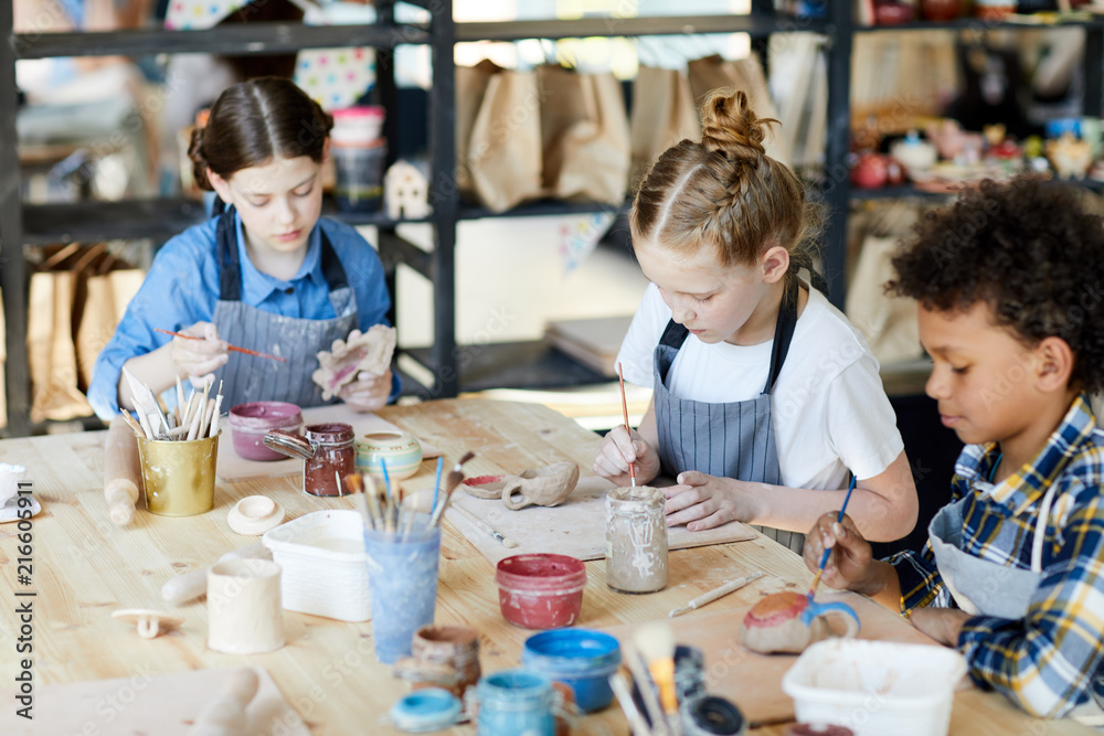 Group of diligent schoolkids sitting by table while making and painting self-made earthenware - obrazy, fototapety, plakaty 