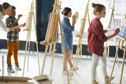 Two schoolgirls and schoolboy standing by their easels and painting while teacher consulting one of them