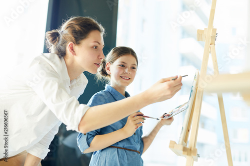 Young teacher of arts pointing at painting of one of pupils while standing by her easel