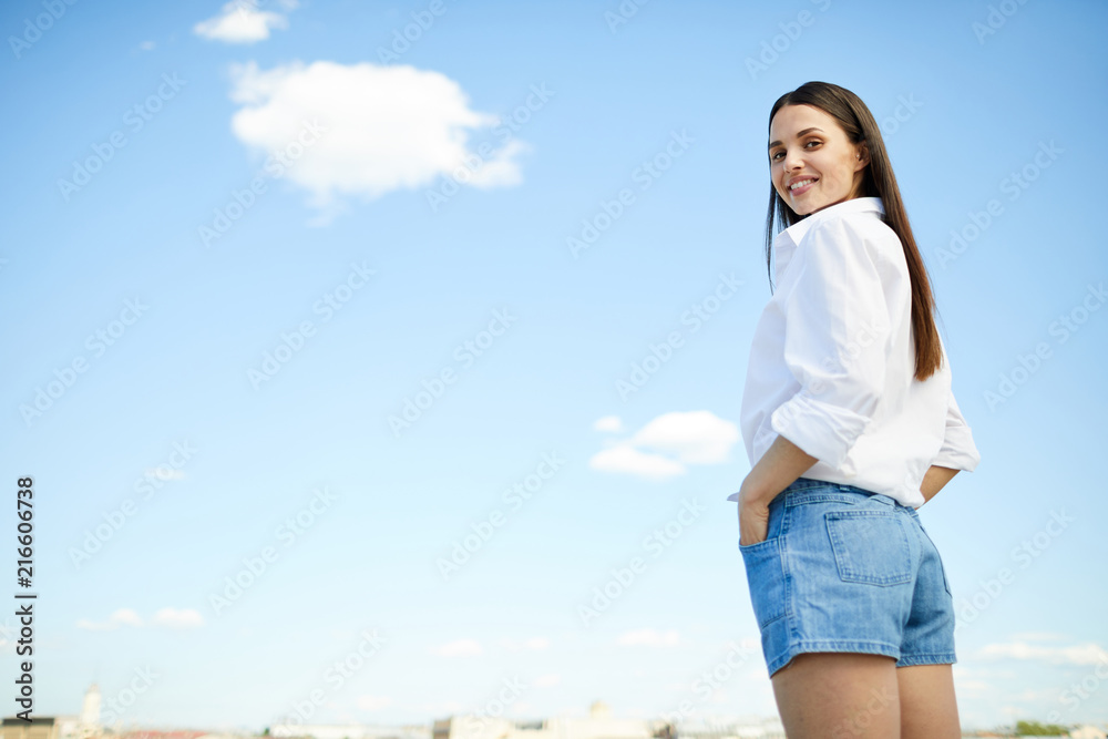 Smiling confident modern girl in casual clothing standing against sky and holding hands in pocket while turning to look at camera Smiling confident modern girl in casual clothing standing against sky and holding hands in pocket while turning to look at camera