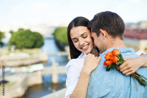 Happy tender young couple hugging each other while dancing on city roof and enjoying romantic date, pretty lady holding small bouquet