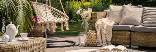 Pillows and blanket on rattan sofa on patio with hanging chair and table during summer. Real photo