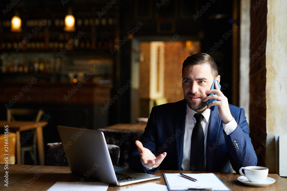 Content confident handsome businessman in formal jacket sitting at restaurant table and solving work issues while talking on mobile phone and gesturing Content confident handsome businessman in formal jacket sitting at restaurant table and solving work issues while talking on mobile phone and gesturing