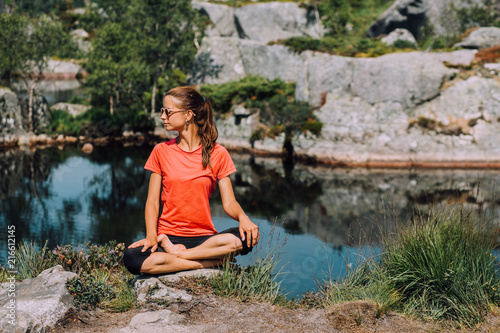 Young girl in pink T-shirt and black leggings sitting on a stone in lotus pose near the lake. concept of yoga  pilates  healthy life.