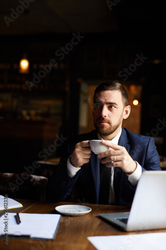 Pensive frowning handsome businessman with beard sitting at table with laptop and drinking hot coffee while waiting for service staff in restaurant