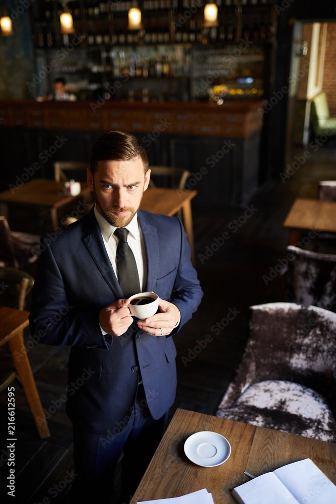 Serious confused young businessman with frowning forehead standing at table and drinking coffee while focusing on thoughts and ideas