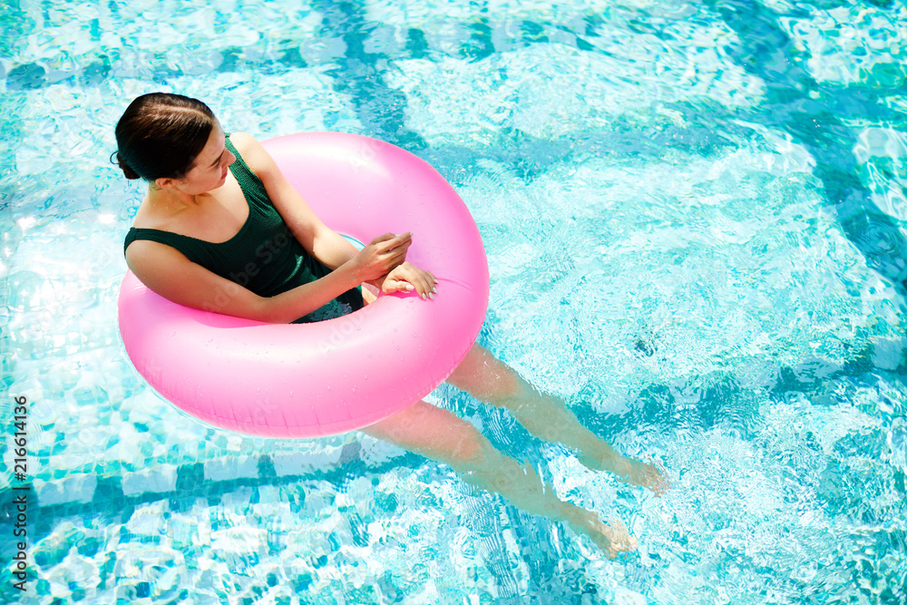 Restful girl inside pink pool buoy balancing on water and enjoying sunny day at spa resort