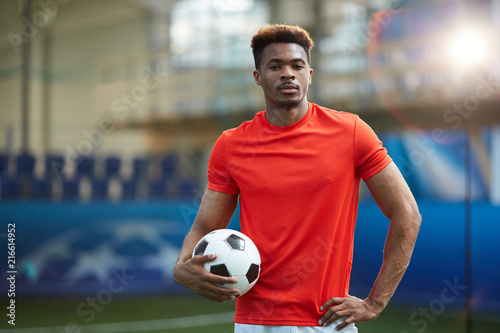 Young African-american man in red football uniform standing with ball at stadium