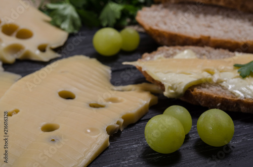 Rural breakfast. A slice of cheese, bread, a sandwich and grapes. on the black wooden table. Close-up image.