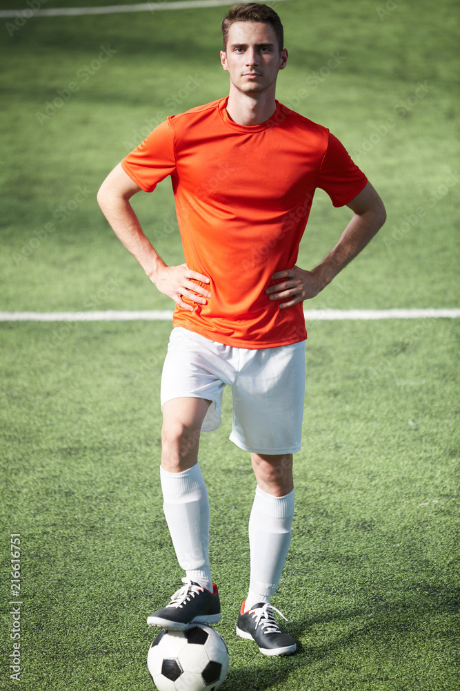 Young football player in uniform standing on green field, keeping his hands on waist and right leg over soccer ball