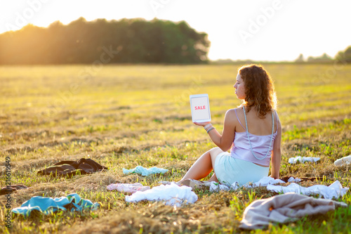 Dressy woman sitting in the middle of the field between scattered cloth and holding a tablet with SALE title during sunset. Fast fashion over-production and pollution concept.