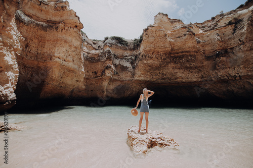 Rear view of young woman in casual clothes standing on rock at the natural beauty cliff beach and enjoy her summer vocation. Back view of woman with hat in hands enjoy the beautiest beach. photo