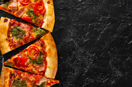 sliced Pizza with Mozzarella cheese, Ham, Tomatoes, pepper, Spices and Fresh Basil pesto sauce. Italian pizza.on black background. with copy space. top view