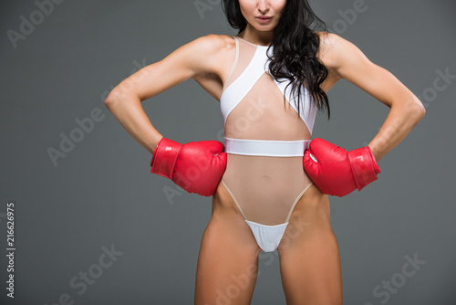 cropped image of sexy sportive woman in white leotard and boxing gloves standing with hands akimbo isolated on grey photo