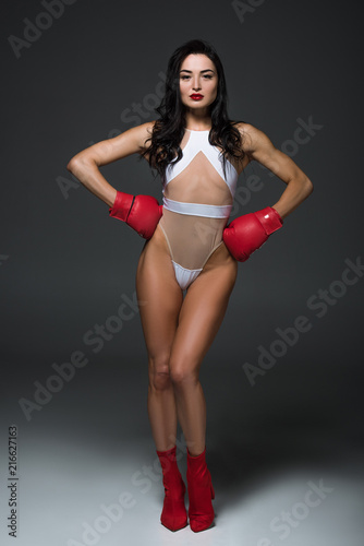 Sexy sportive woman in white swimwear and boxing gloves standing with hands akimbo, on grey