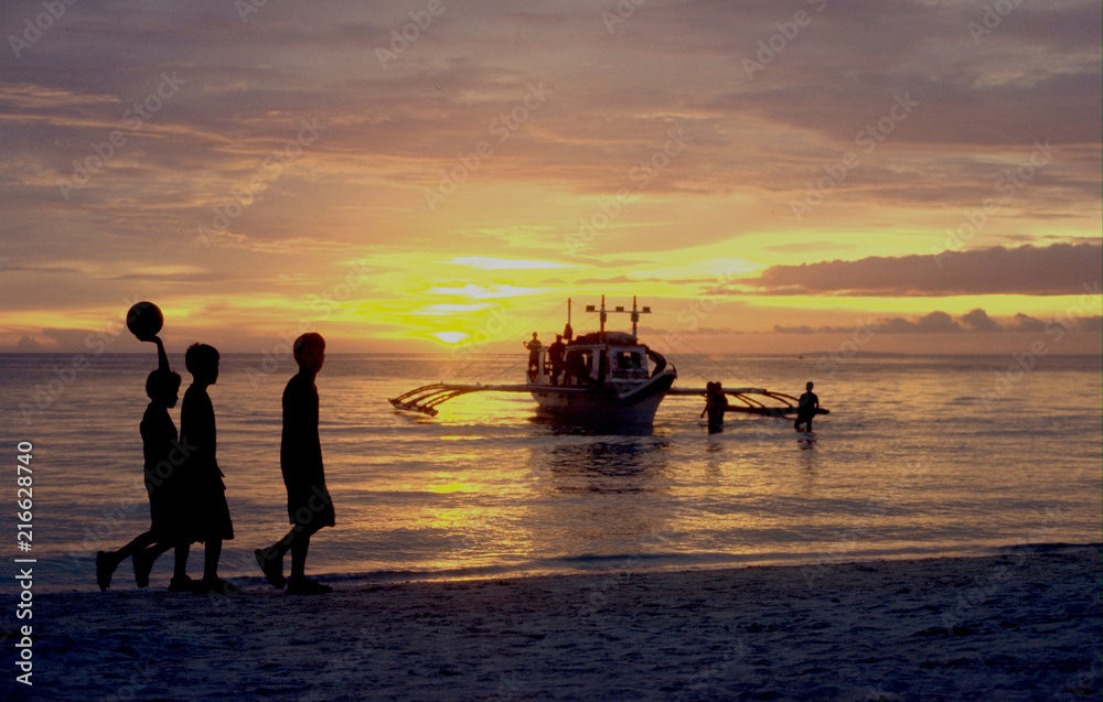 Philipppines: Football boys at sunset walking along Boracay Beach and a boat is arriving at the Island too