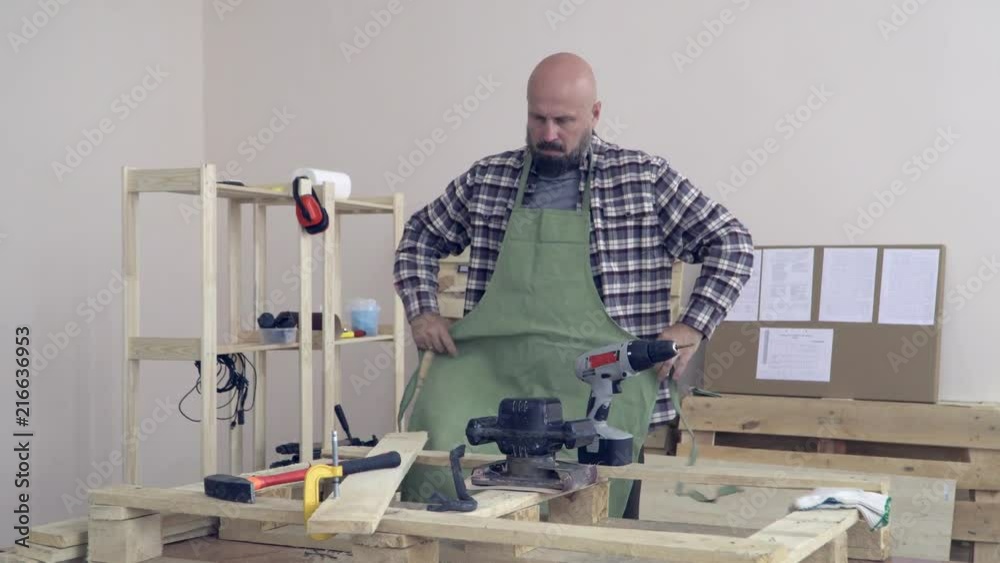 An adult bearded bald carpenter wears a green apron and rolls up the sleeves of a plaid shirt near the workbench. Against the carpenter's workshop, a Board with instructions and racks of tools. An adult bearded bald carpenter wears a green apron and rolls up the sleeves of a plaid shirt near the workbench. Against the carpenter's workshop, a Board with instructions and racks of tools.