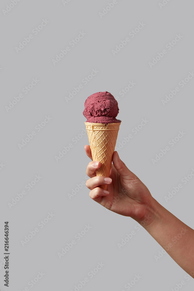 A female hand holds red berry ice cream in a waffle cone on a light gray background. A female hand holds red berry ice cream in a waffle cone on a light gray background.