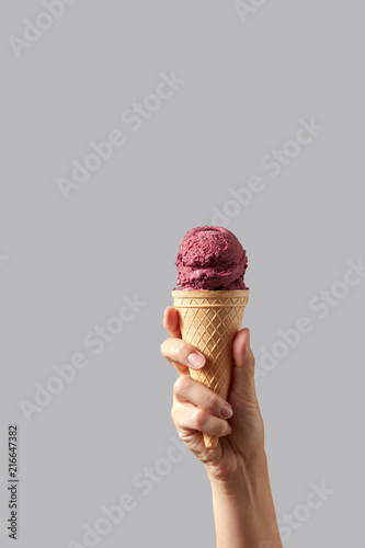 A woman's hand holds a waffle cone with red cherry sorbet on a light gray background.