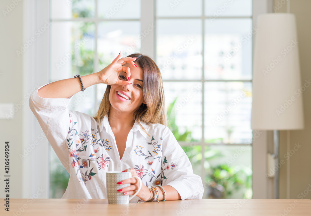 Young woman at home drinking coffee with happy face smiling doing ok sign with hand on eye looking through fingers Young woman at home drinking coffee with happy face smiling doing ok sign with hand on eye looking through fingers