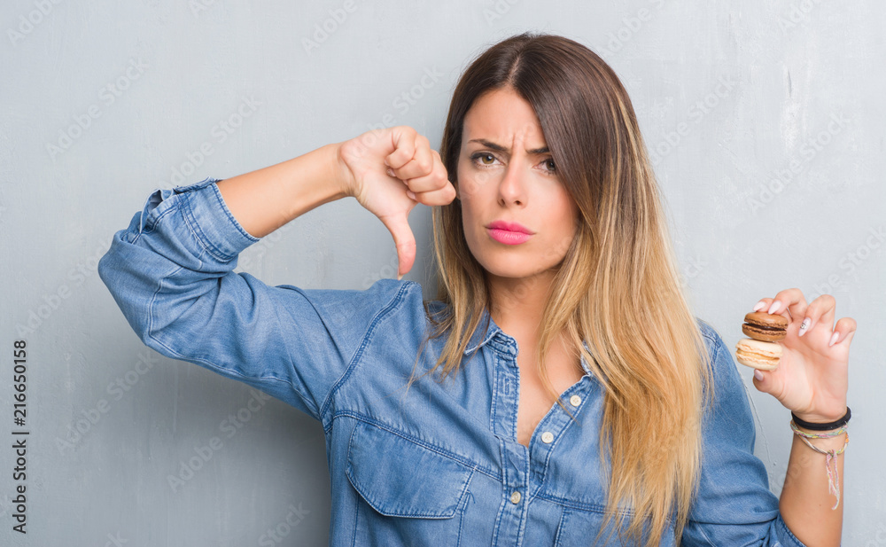 Fototapeta premium Young adult woman over grey grunge wall eating chocolate macarons with angry face, negative sign showing dislike with thumbs down, rejection concept
