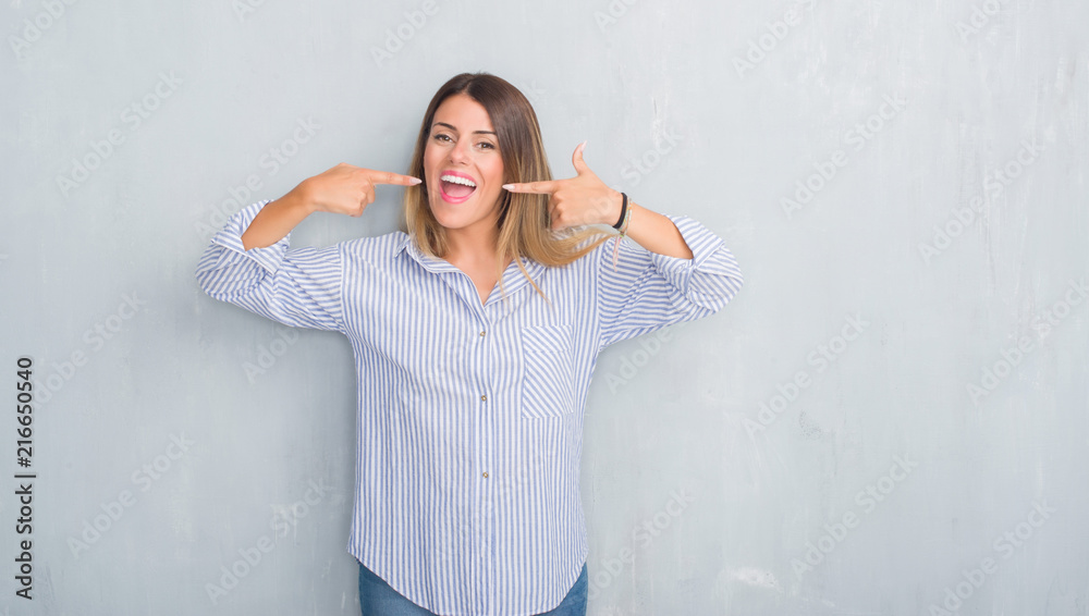 Young adult woman over grey grunge wall wearing fashion business outfit smiling confident showing and pointing with fingers teeth and mouth. Health concept.