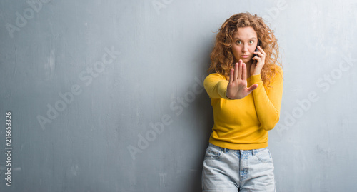 Young redhead woman over grey grunge wall talking on the phone with open hand doing stop sign with serious and confident expression, defense gesture