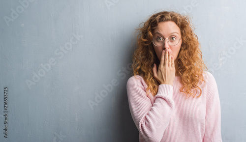 Young redhead woman over grey grunge wall wearing glasses cover mouth with hand shocked with shame for mistake, expression of fear, scared in silence, secret concept