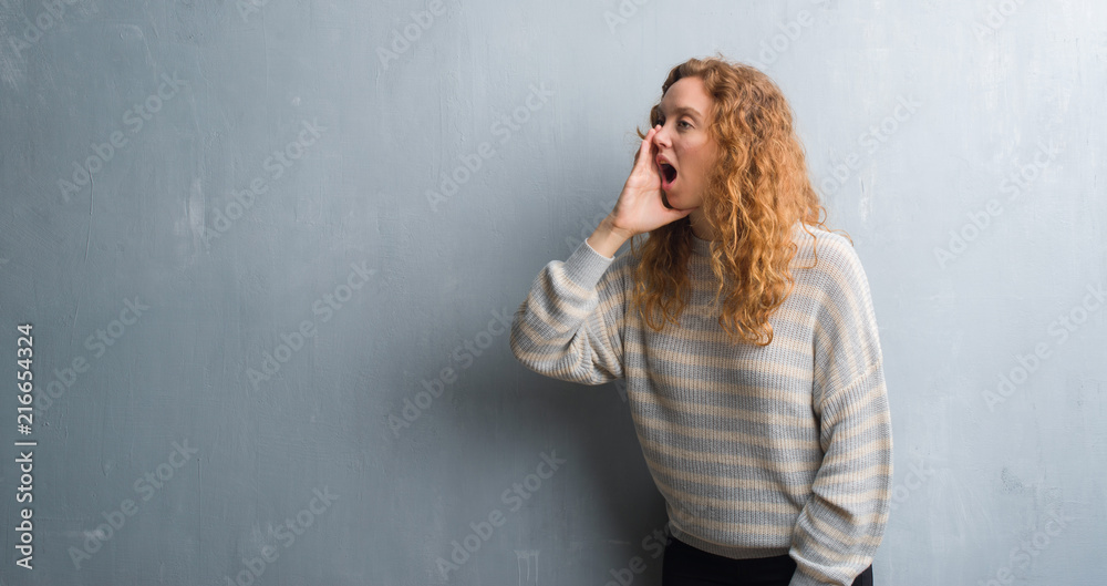 Young redhead woman over grey grunge wall shouting and screaming loud to side with hand on mouth. Communication concept. Young redhead woman over grey grunge wall shouting and screaming loud to side with hand on mouth. Communication concept.