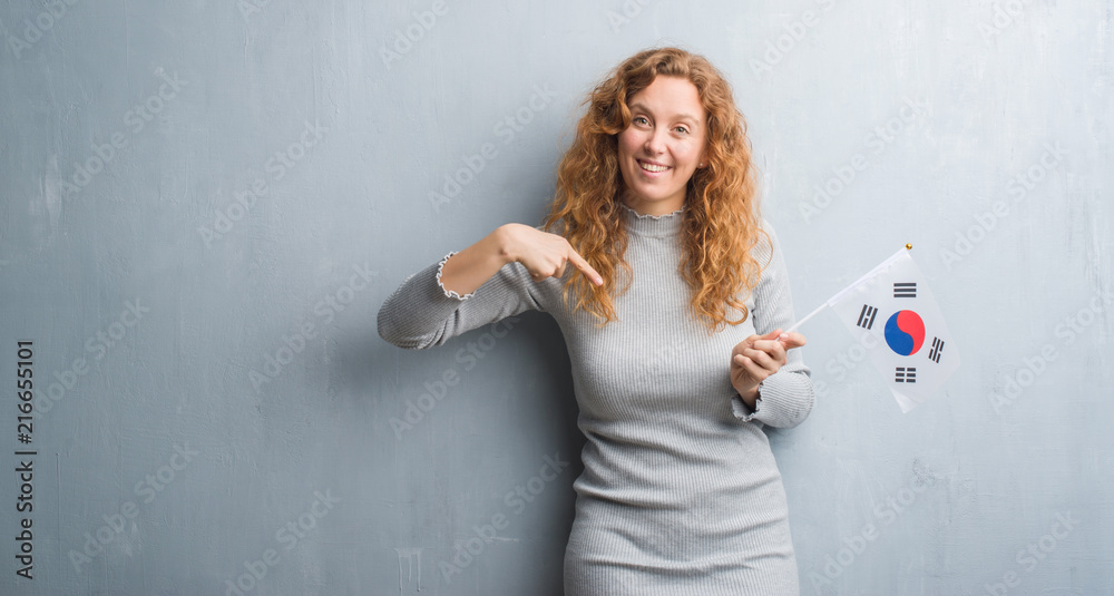 Young redhead woman over grey grunge wall holding flag of South Korea with surprise face pointing finger to himself Young redhead woman over grey grunge wall holding flag of South Korea with surprise face pointing finger to himself