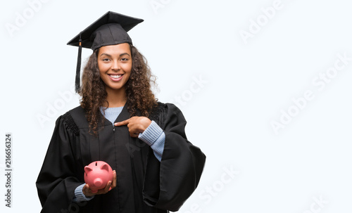 Young hispanic woman wearing graduation uniform holding piggy bank with surprise face pointing finger to himself