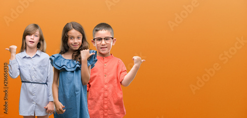 Group of boy and girls kids over orange background pointing and showing with thumb up to the side with happy face smiling