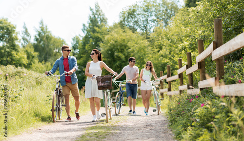 people, leisure and lifestyle concept - happy young friends with fixed gear bicycles walking along country road in summer