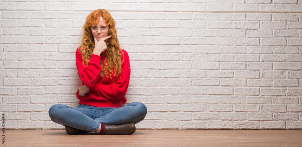 Young redhead woman sitting over brick wall looking confident at the camera with smile with crossed arms and hand raised on chin. Thinking positive. Young redhead woman sitting over brick wall looking confident at the camera with smile with crossed arms and hand raised on chin. Thinking positive.