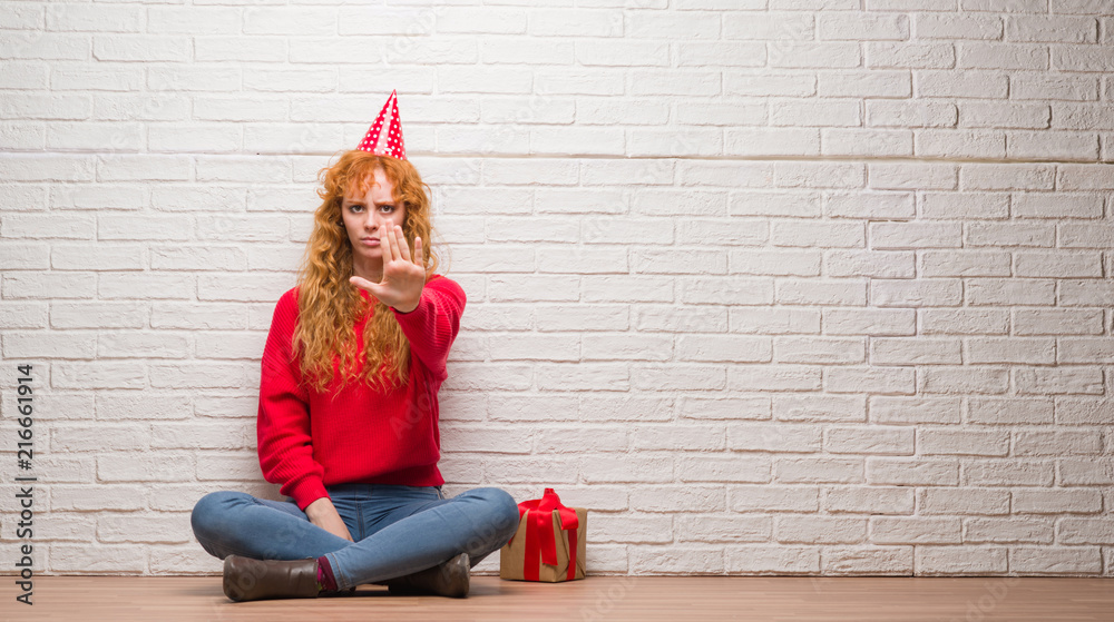 Young redhead woman sitting over brick wall wearing birthday hat with open hand doing stop sign with serious and confident expression, defense gesture Young redhead woman sitting over brick wall wearing birthday hat with open hand doing stop sign with serious and confident expression, defense gesture