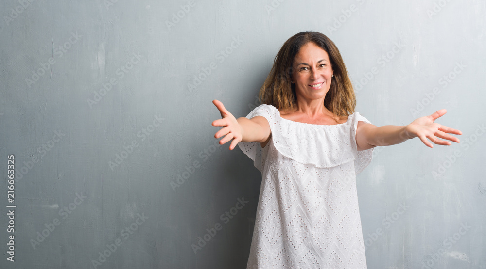 Middle age hispanic woman standing over grey grunge wall looking at the camera smiling with open arms for hug. Cheerful expression embracing happiness. Middle age hispanic woman standing over grey grunge wall looking at the camera smiling with open arms for hug. Cheerful expression embracing happiness.