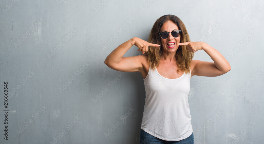 Middle age hispanic woman over grey wall wearing sunglasses smiling confident showing and pointing with fingers teeth and mouth. Health concept.