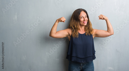 Middle age hispanic woman standing over grey grunge wall showing arms muscles smiling proud. Fitness concept.