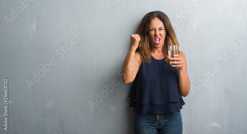 Middle age hispanic woman drinking glass of water annoyed and frustrated shouting with anger, crazy and yelling with raised hand, anger concept