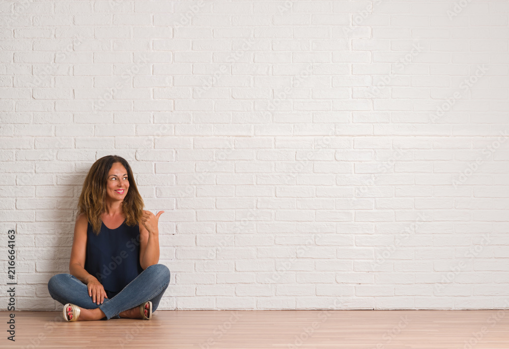 Middle age hispanic woman sitting on the floor over white brick wall smiling with happy face looking and pointing to the side with thumb up. Middle age hispanic woman sitting on the floor over white brick wall smiling with happy face looking and pointing to the side with thumb up.
