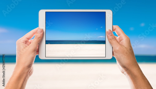 Hands holding tablet with holiday beach and sea on screen and beach and coast background