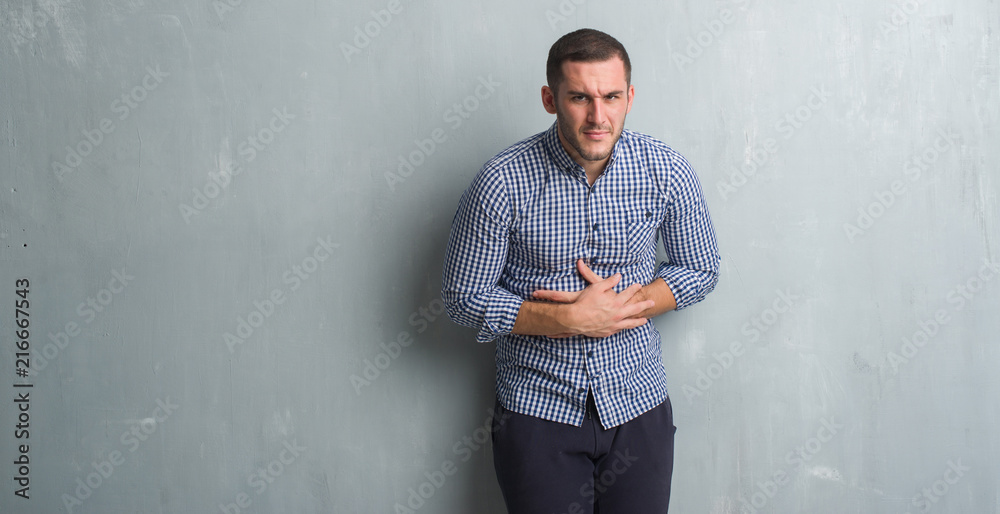 Young caucasian man over grey grunge wall with hand on stomach because nausea, painful disease feeling unwell. Ache concept.