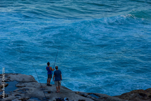 Two tourists standing with fishing rod in hand on the edge of rock cliff at Tamarama beach