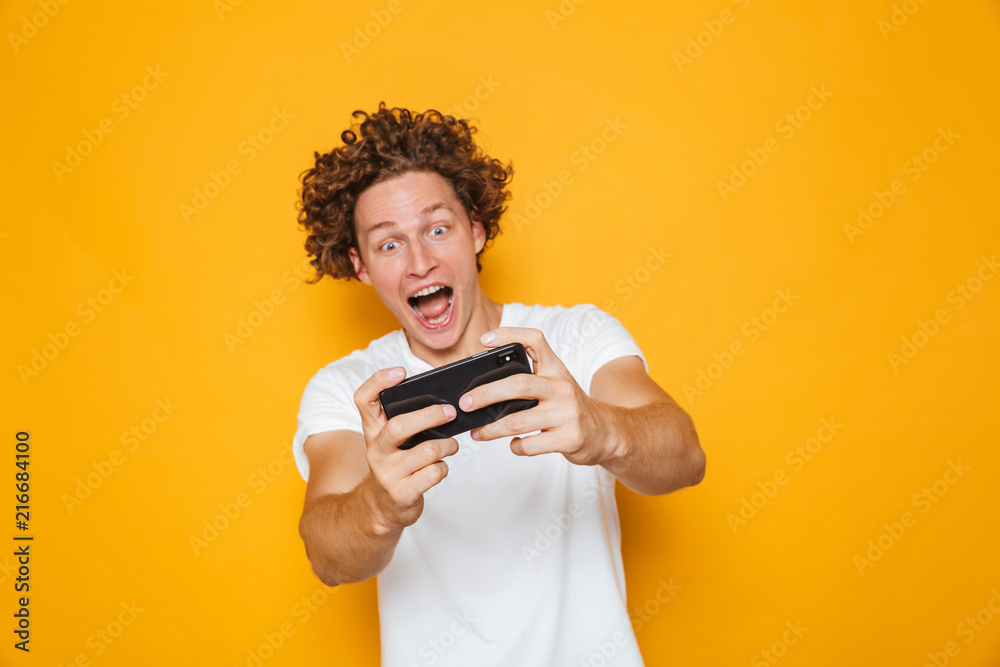 Cheerful excited guy in casual t-shirt playing online video games using mobile phone, isolated over yellow background Cheerful excited guy in casual t-shirt playing online video games using mobile phone, isolated over yellow background