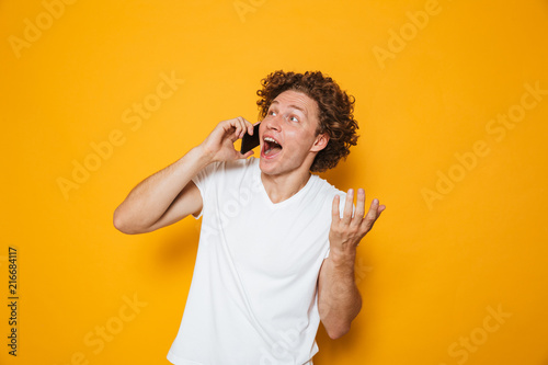 Photo of happy young guy with curly hair shouting and talking on smartphone  isolated over yellow background