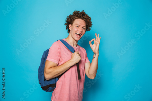 Image of student guy with curly hair wearing casual clothing and backpack smiling and showing ok sign, isolated over blue background