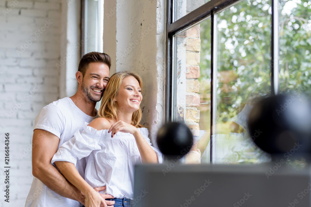 A beautiful young woman standing next to the window with boyfriend hugging her from behind. A beautiful young woman standing next to the window with boyfriend hugging her from behind.