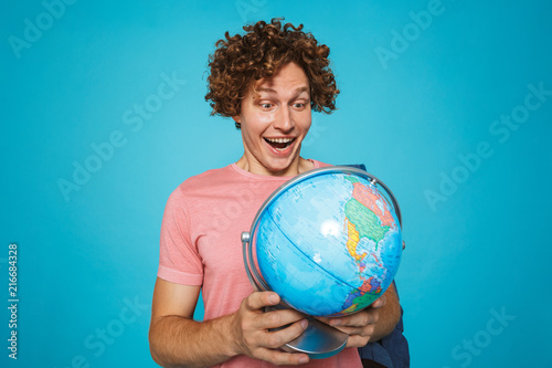 Photo of teenage guy with curly hair wearing backpack smiling and holding earth globe, isolated over blue background