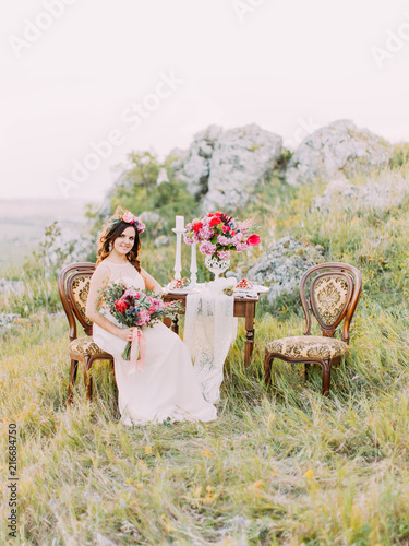 The beautiful bride with the bouwuet is sitting on the antique chair near the wedding table in the mountains. photo